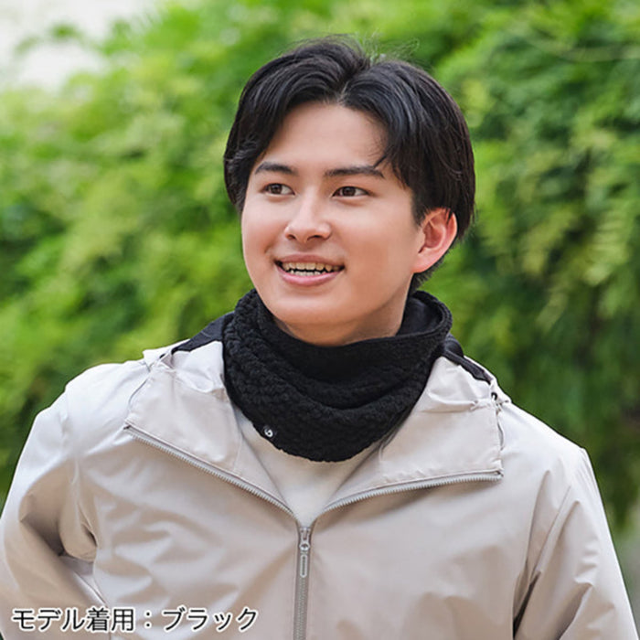 A man wearing a beige zip-up jacket and a black knit neck warmer, photographed outdoors with a blurred green foliage background